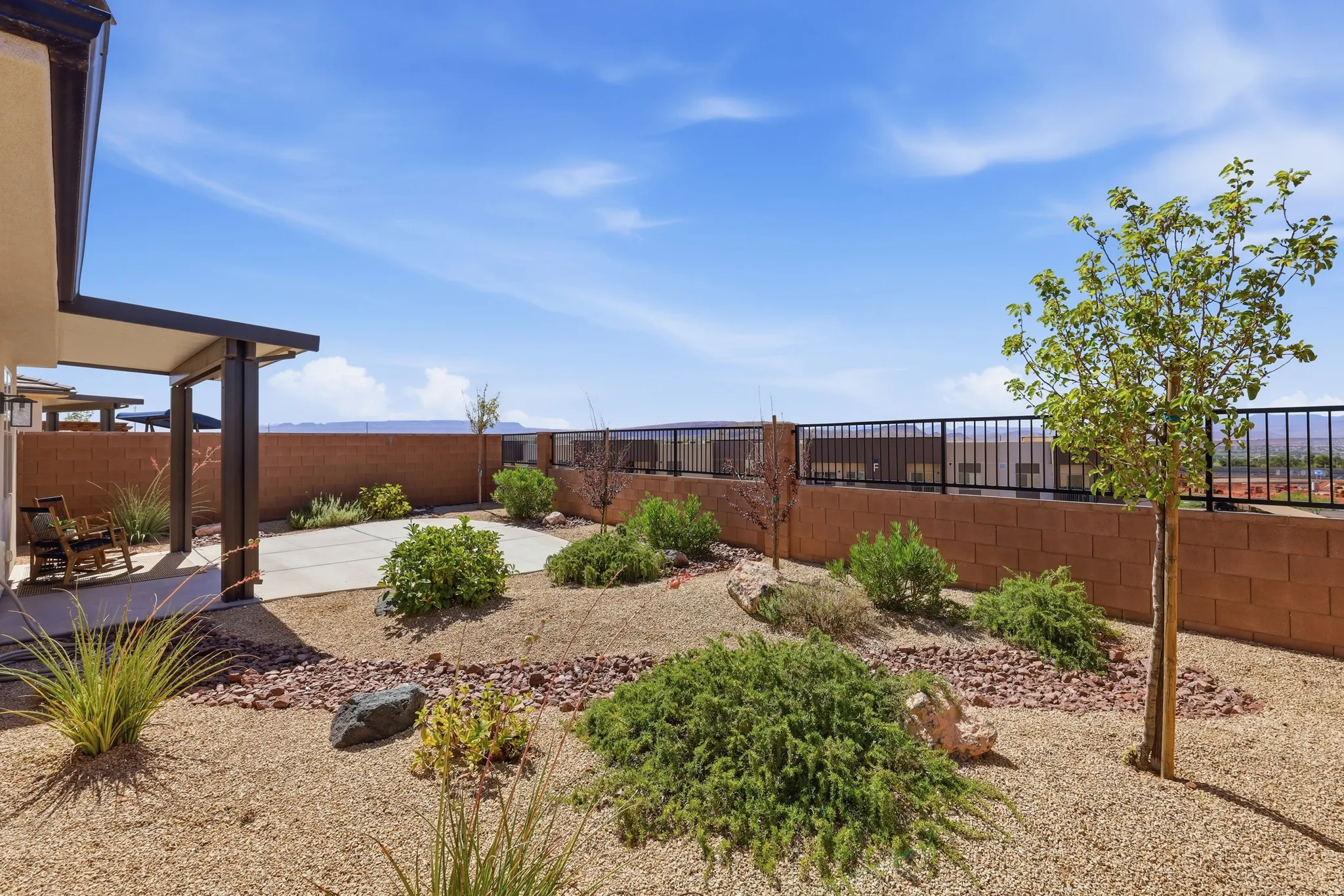 Expansive backyard featuring a covered patio with concrete flooring, a decorative rock landscape, a block wall perimeter, a wrought iron fence, and established shrubs and young trees