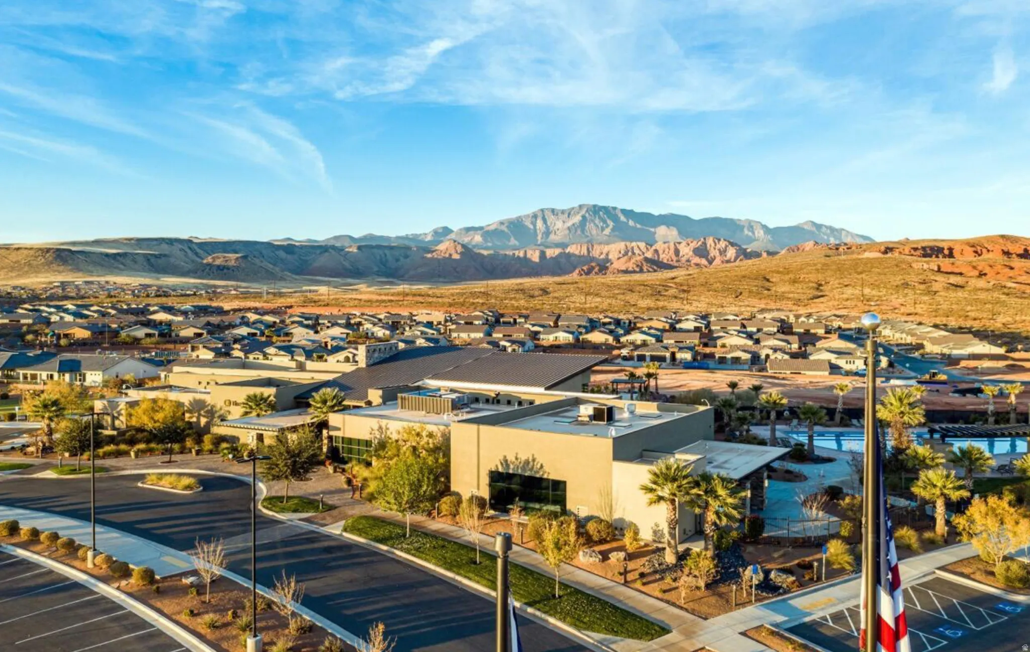 Expansive community featuring desert landscaping, paved roadways, multiple buildings with large windows, and a mountain range backdrop