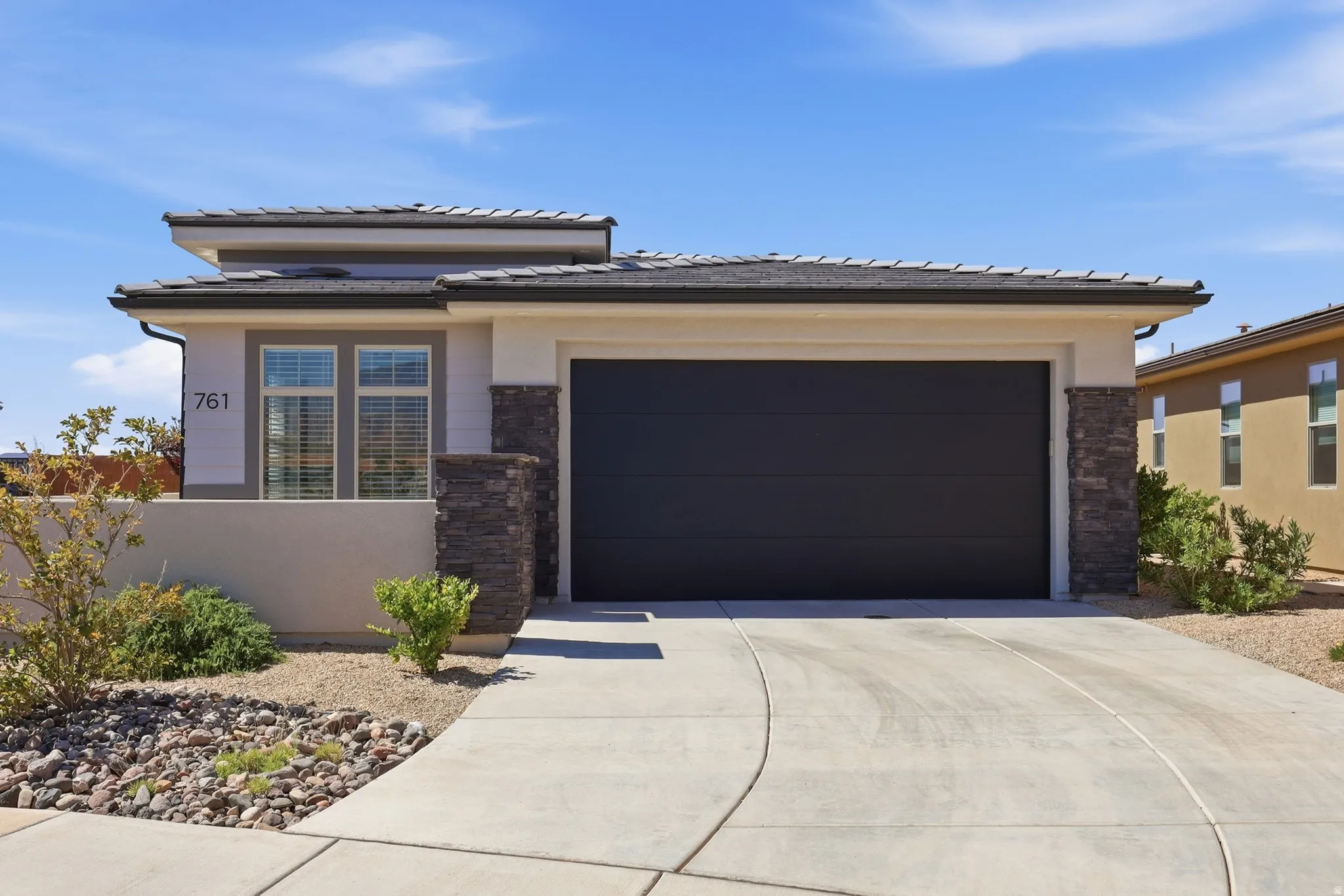 Contemporary stucco exterior with a dark garage door, stone pillar accents, and a low-maintenance landscape design