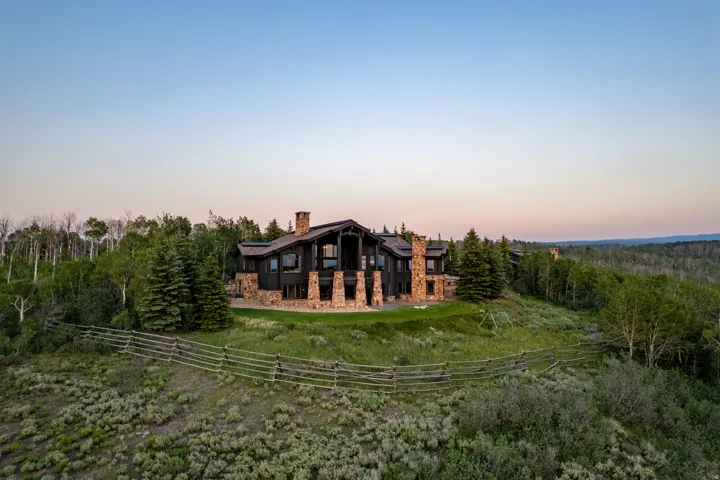 Expansive property featuring a multi-level structure, stone foundation and chimneys, dark siding, multiple windows, and natural landscaping with mature trees and a rustic fence