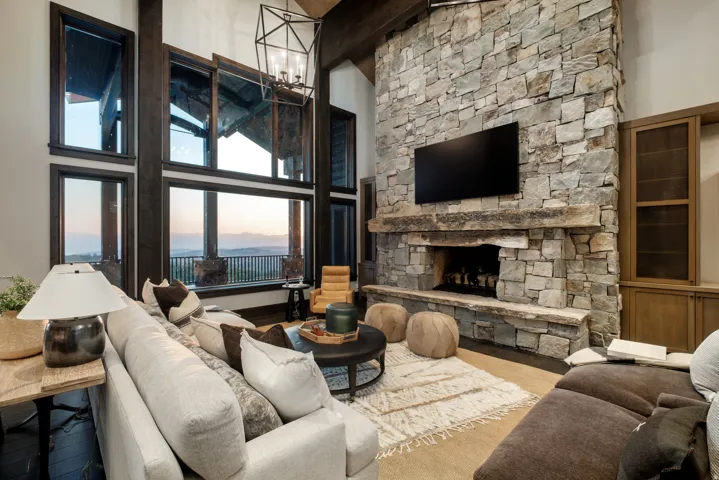 Floor-to-ceiling windows with dark wood framing, a stone-clad fireplace with a natural wood mantel, built-in wooden cabinetry, and exposed ceiling beams