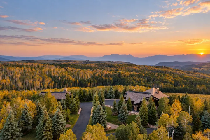 Extensive natural landscaping with mature trees, a private paved driveway, and multiple residential structures, all set against a mountainous backdrop