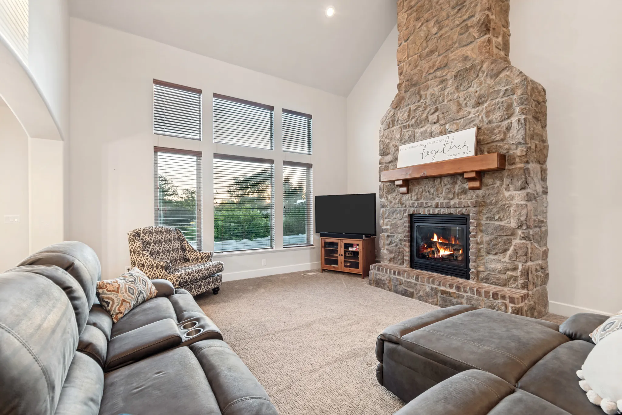 Great room featuring vaulted ceilings, a stone fireplace with a wood mantle, and large windows providing ample amounts of natural light.