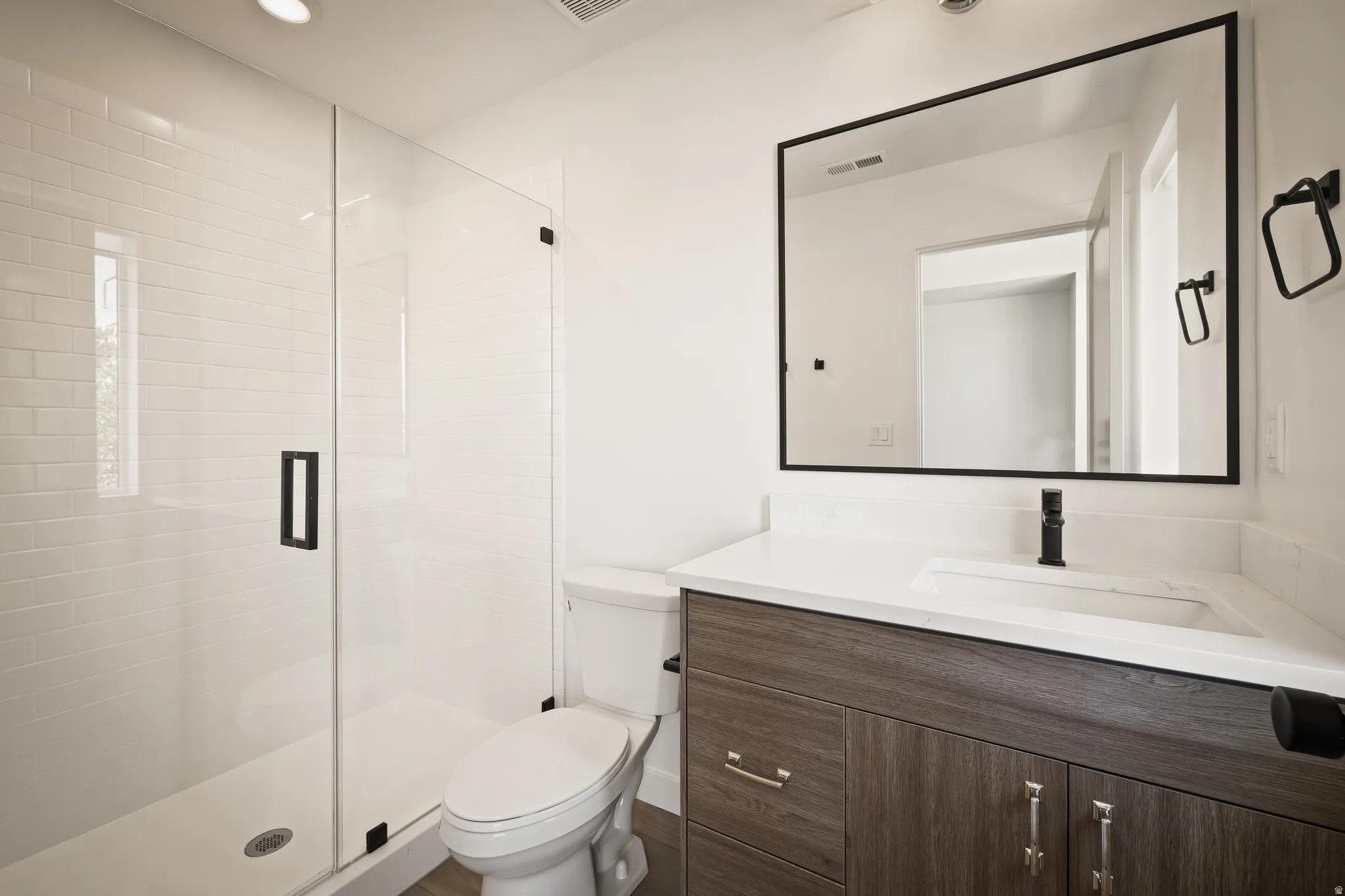 Bathroom featuring a glass-enclosed shower with white subway tile, a toilet, a wood-finish vanity with an integrated sink, a white countertop, and a rectangular black-framed mirror
