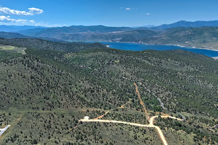 Expansive mountain landscape featuring a prominent blue lake, rolling hills covered in evergreen trees, and visible dirt roads