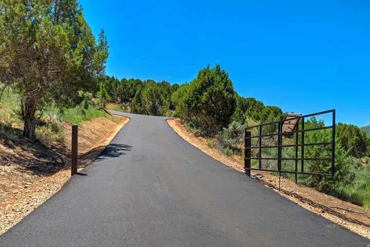 Paved driveway leading through a natural, treed landscape with a metal gate