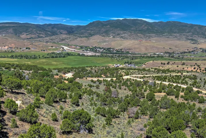 Expansive mountain and valley scenery featuring rolling hillsides, green fields, distant structures, and a clear blue sky