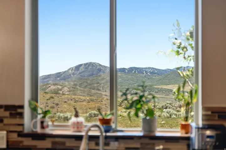 Bright kitchen featuring expansive windows framing panoramic mountain and valley vistas, natural light flooding the workspace, potted greenery on the sill, and a stainless steel sink with modern fixtures overlooking the landscape