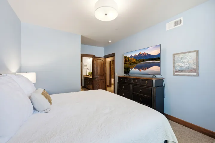 Serene primary bedroom showcasing soft blue walls, recessed ceiling lighting, wood-finish flooring with carpet accents, dark wood dresser with ample storage, and framed landscape artwork