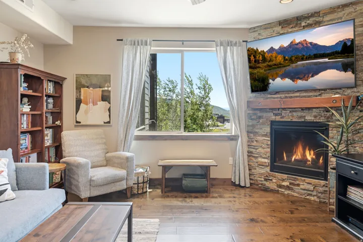 Spacious living area featuring a gas fireplace with stone surround, wood-finish flooring, and expansive windows framed with white curtains