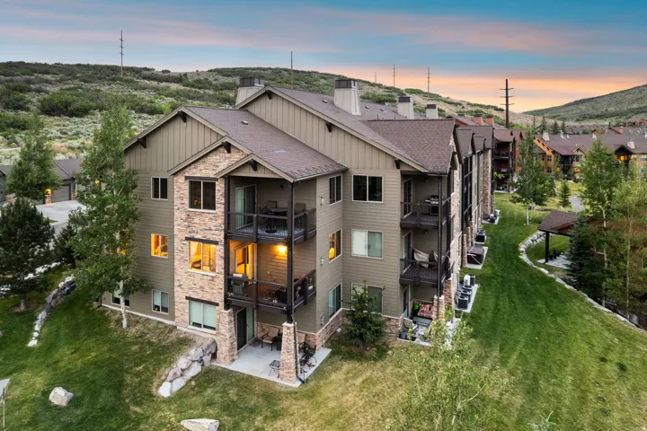 Modern multi-level residence showcasing mixed material exterior with stone and metal siding, expansive windows with evening illumination, manicured lawn with mature landscaping, and hillside setting with panoramic backdrop