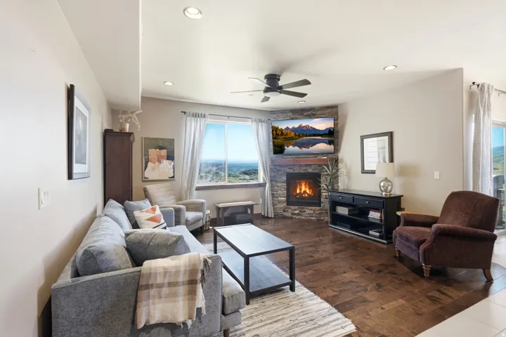 Living area featuring wood-finish flooring, a stone-surround fireplace, a ceiling fan with integrated lighting, recessed lighting, and expansive windows