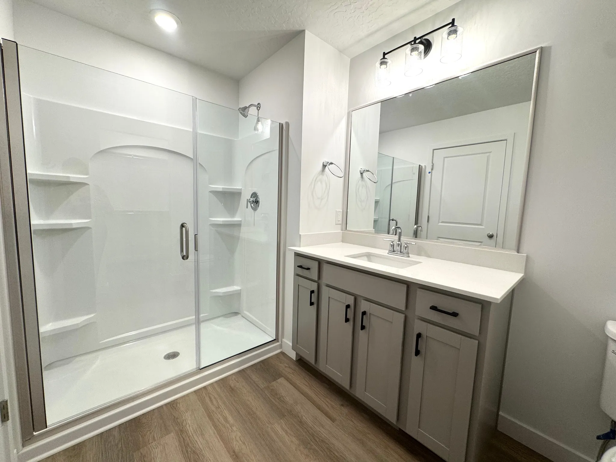 Primary  bathroom featuring a glass-enclosed shower with integrated shelving, a single vanity with a white countertop, wood-finish flooring, and overhead vanity lighting