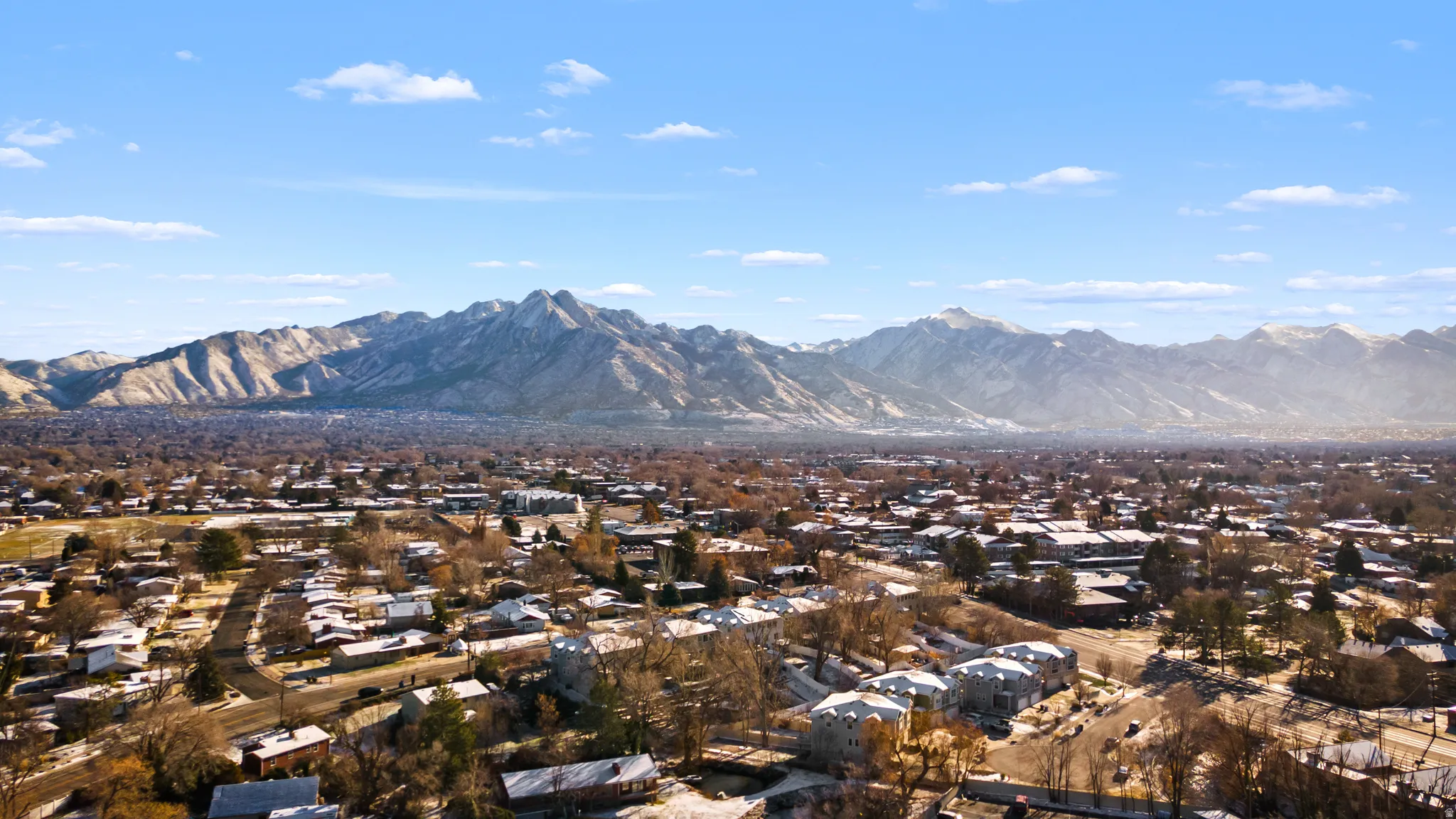 Expansive aerial perspective showcasing a residential area with varied housing structures, mature trees, and mountainous terrain in the distance