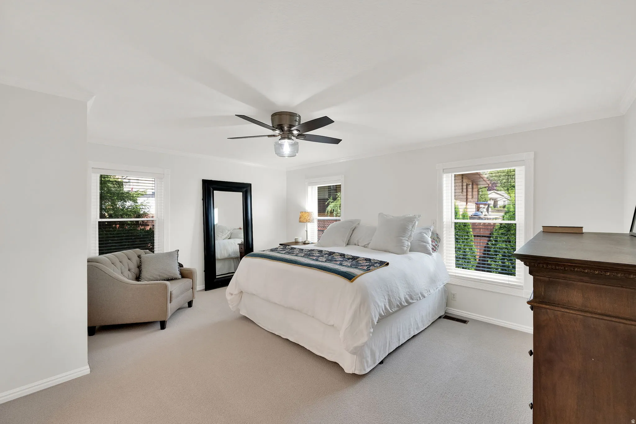 Spacious primary suit featuring light-colored carpeting, multiple windows with blinds, a ceiling fan with integrated lighting, and crown molding