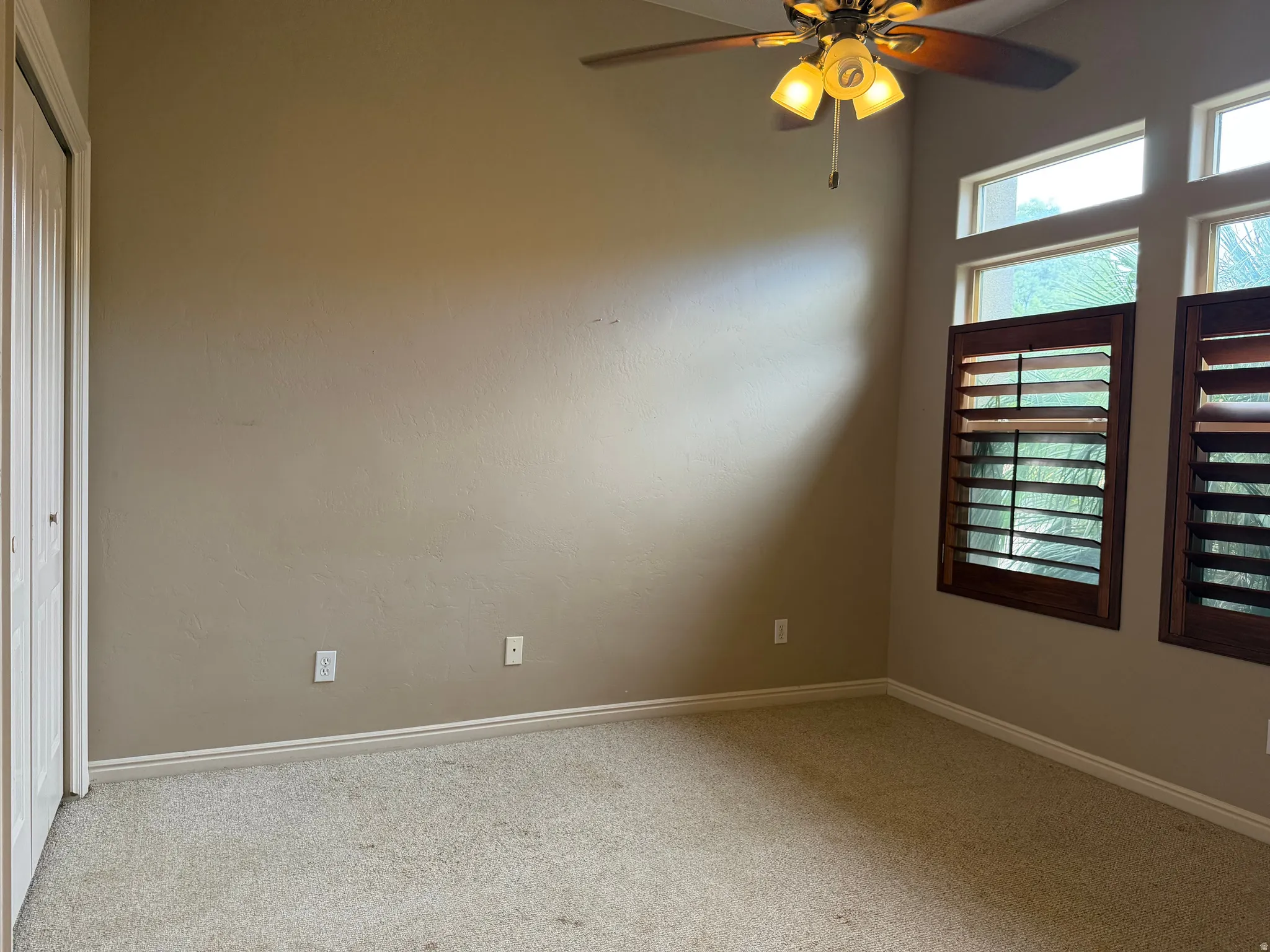 Carpeted room featuring a ceiling fan with integrated lighting, multiple windows with wood plantation shutters, and a bi-fold closet door