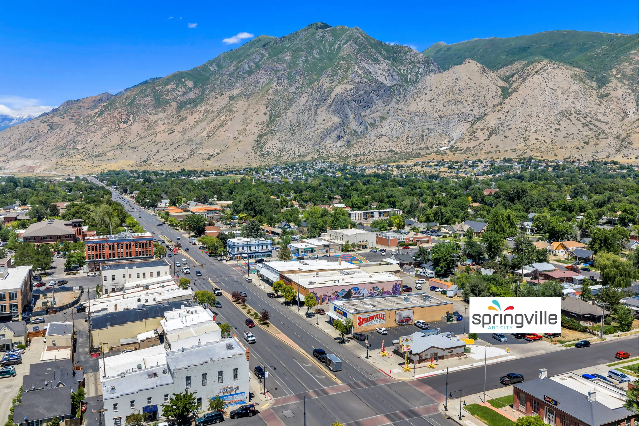 Expansive aerial perspective showcasing a bustling commercial thoroughfare with multiple buildings, a broad central road, a mountainous backdrop, and dense surrounding greenery