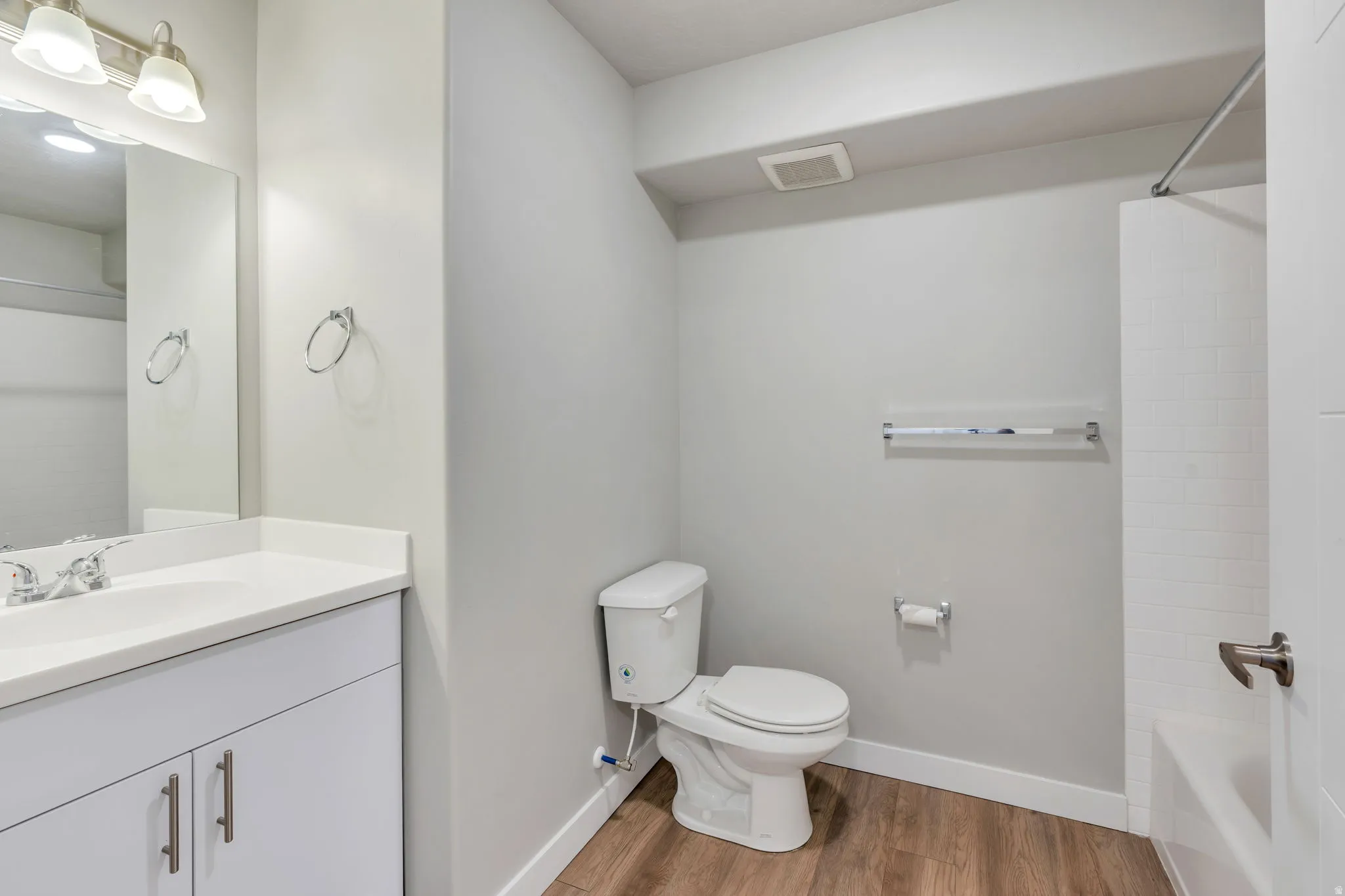 Bathroom featuring a white vanity with a light-colored countertop, chrome fixtures, a toilet, a bathtub with white subway tile surround, and wood-finish flooring
