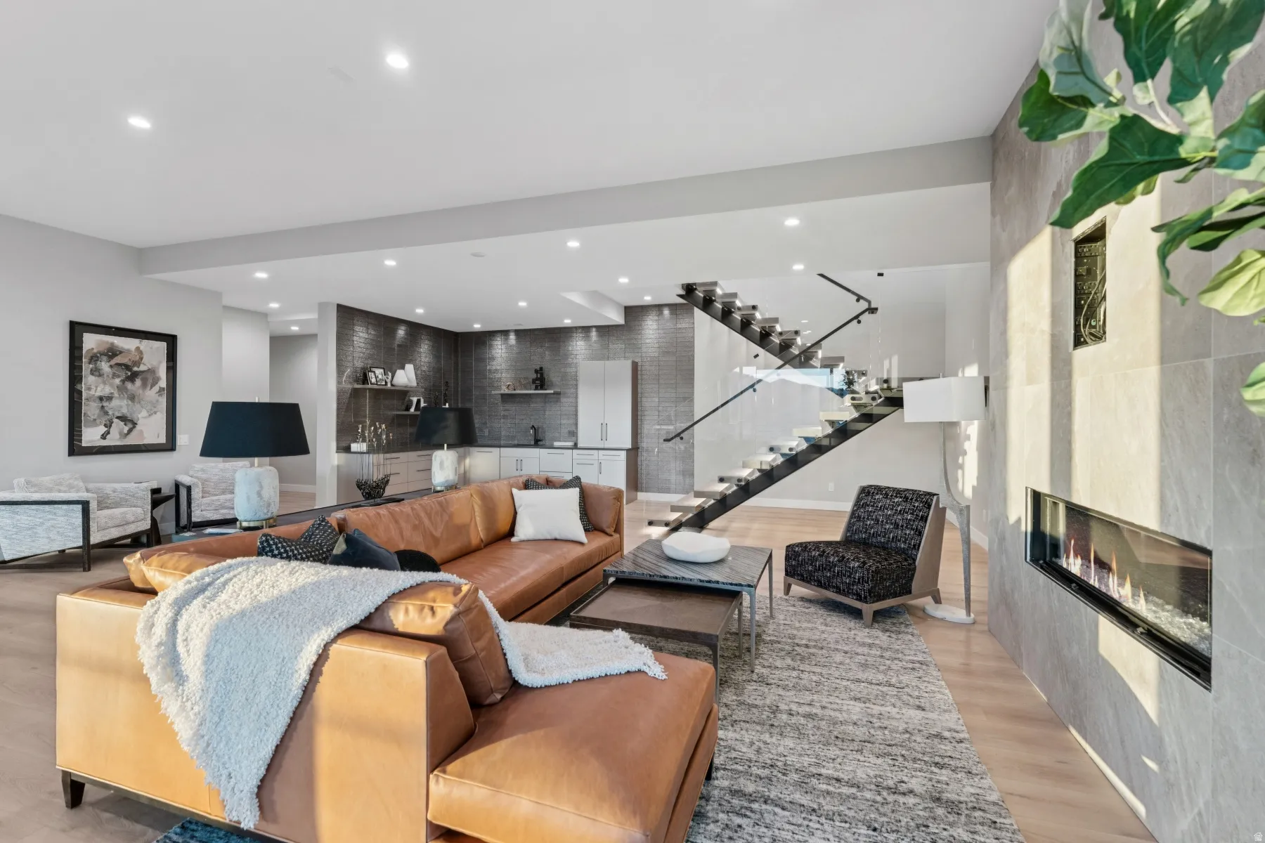 Open-concept living area featuring light wood-finish flooring, a contemporary floating staircase with glass railing, and a linear fireplace inset into a large format tile wall