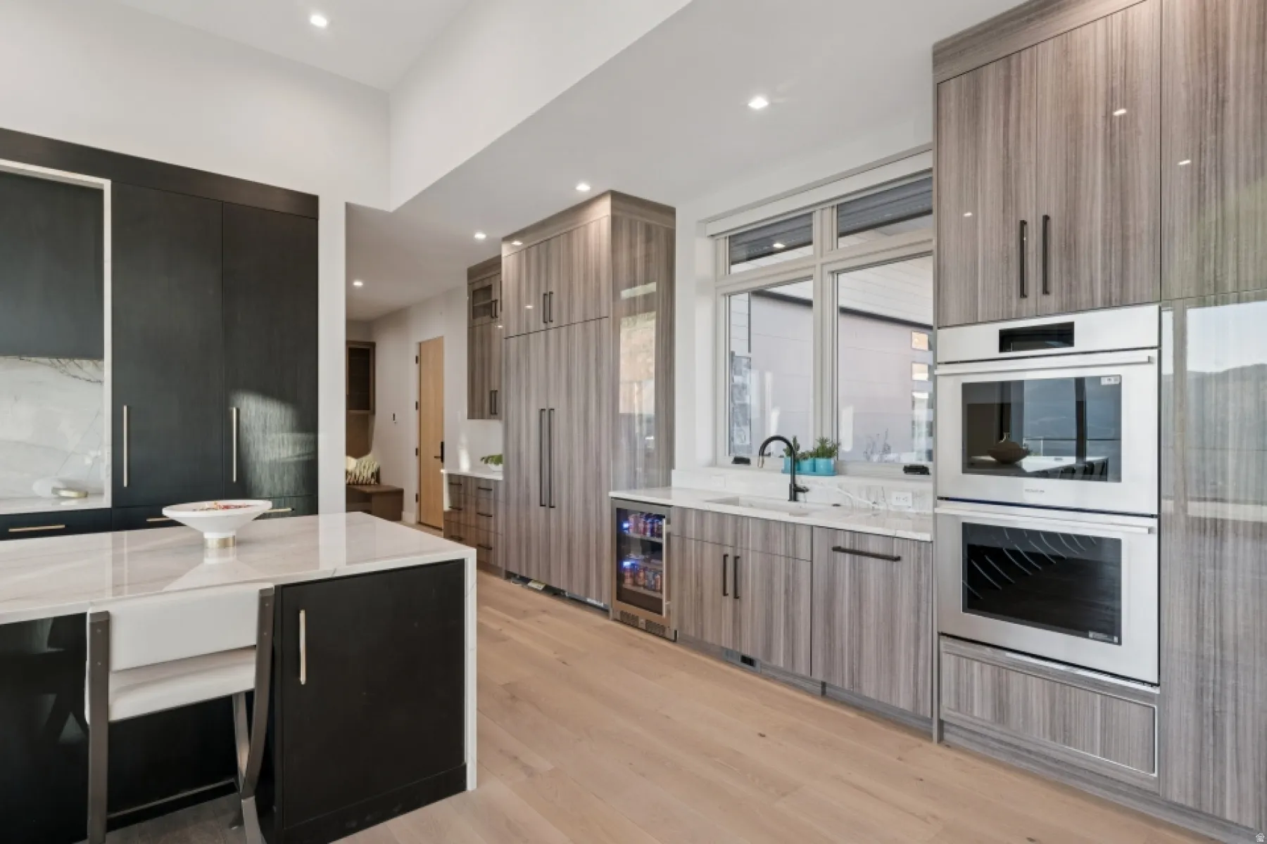 Gourmet kitchen featuring light wood-finish flooring, a central island with a farmhouse sink, integrated stainless steel appliances, a beverage refrigerator, and light wood-finish cabinetry
