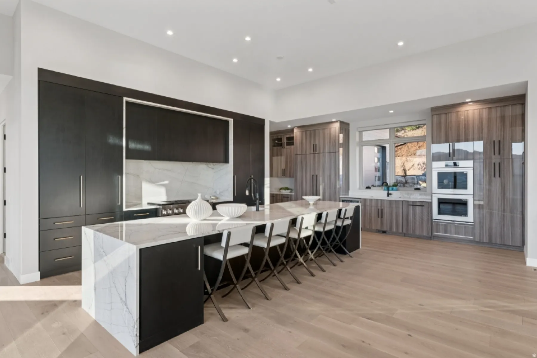 Gourmet kitchen featuring a large central island with waterfall countertop, integrated cooktop, dual wall ovens, and custom cabinetry with varied wood-grain finishes