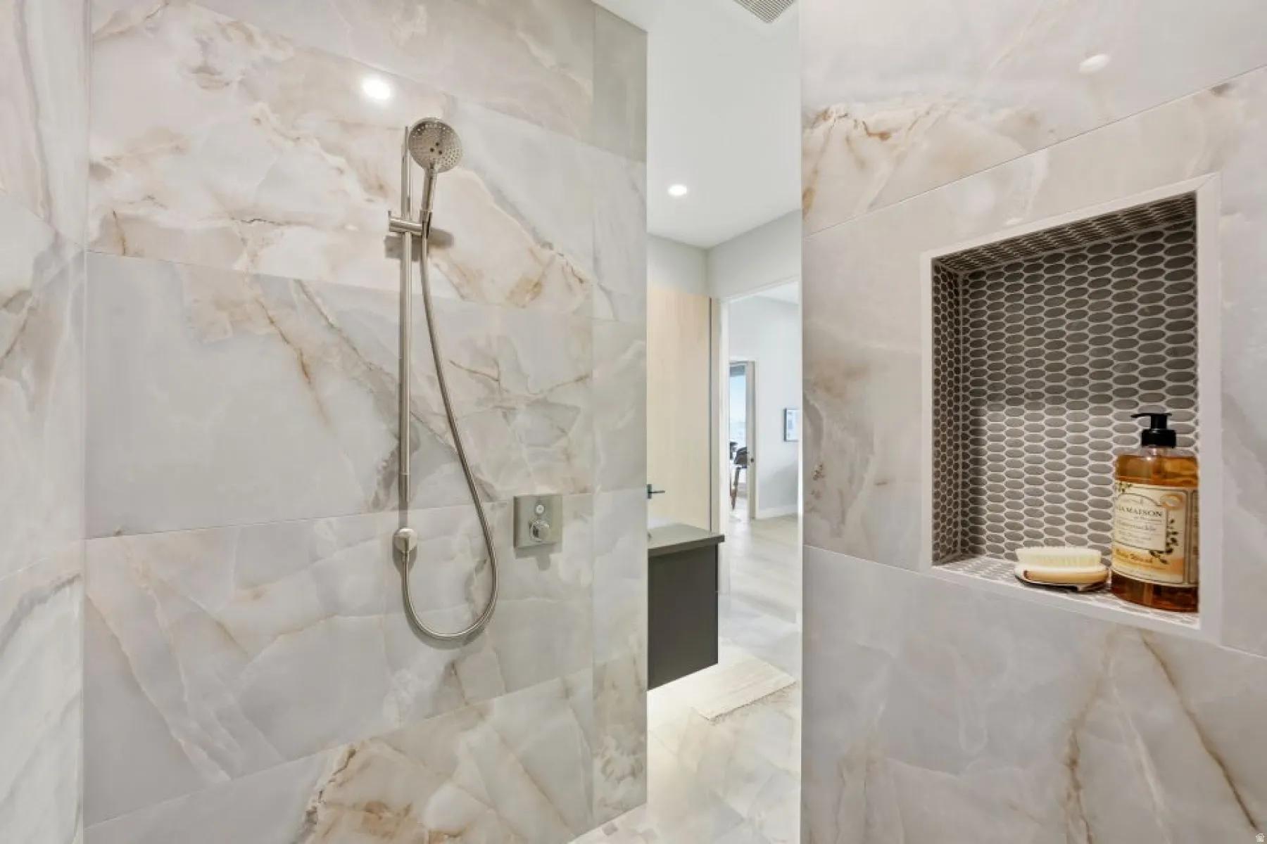Spacious shower enclosure featuring large-format veined wall tiles, a built-in niche with mosaic tile backing, and a brushed nickel showerhead with a handheld sprayer