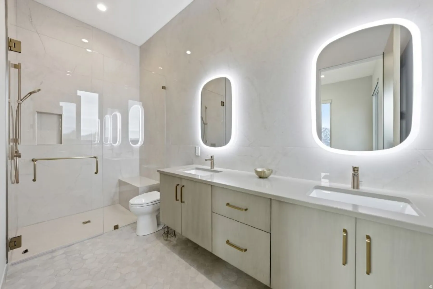 Dual vanity with integrated sinks, back-lit mirrors, a glass-enclosed shower, a built-in shower bench, and hexagonal tile flooring