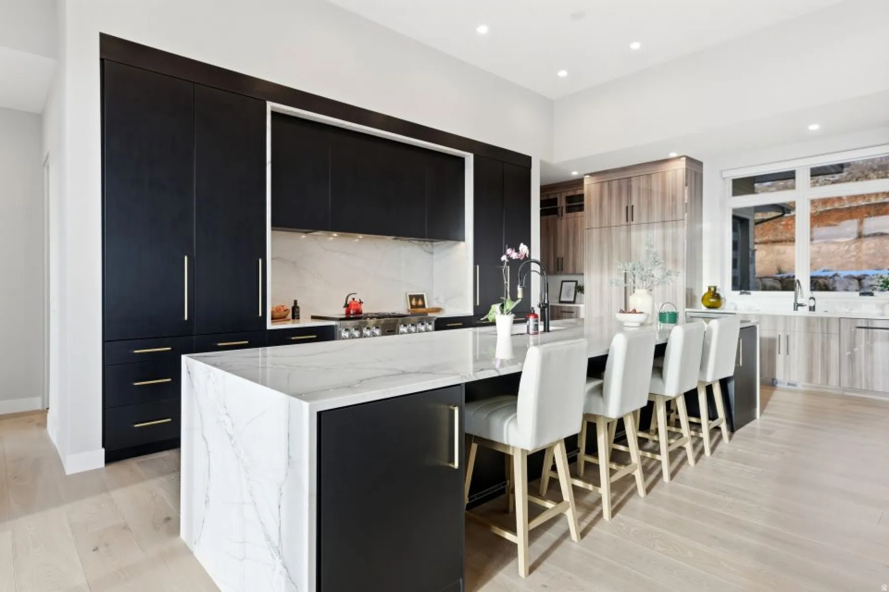Modern kitchen featuring a waterfall island with a stone surface, contrasting dark cabinetry, recessed lighting, wood-finish flooring, and a large window