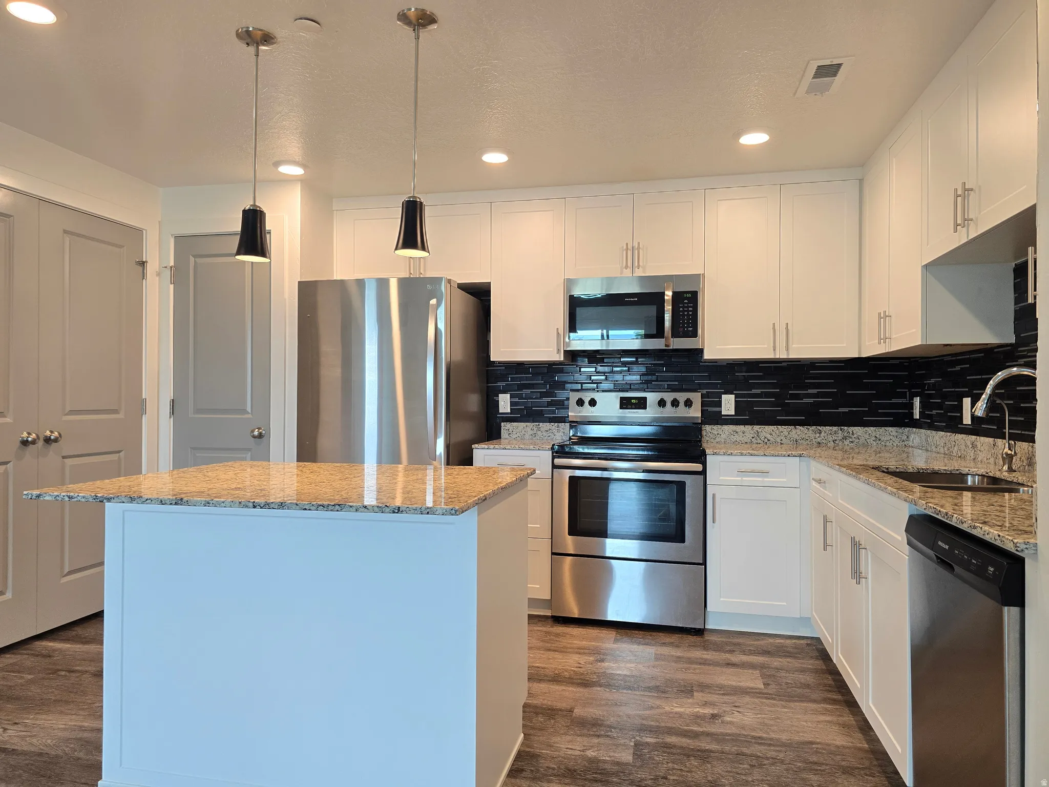 Kitchen featuring a central island with a granite countertop, stainless steel appliances, white cabinetry, a dark tile backsplash, and wood-finish flooring