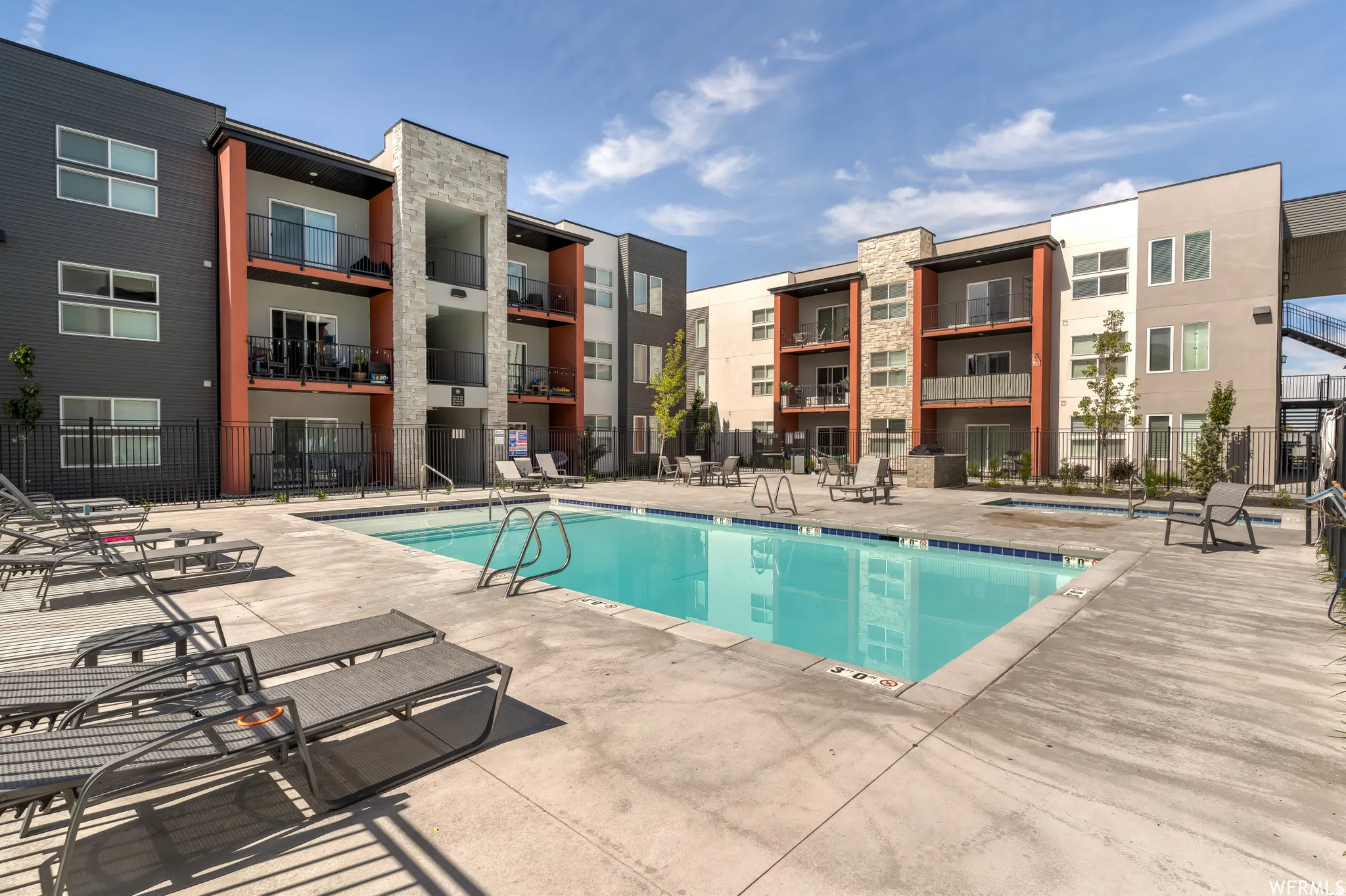 Outdoor swimming pool with integrated concrete deck, surrounded by contemporary multi-story buildings featuring balconies, dark siding, and stone accents