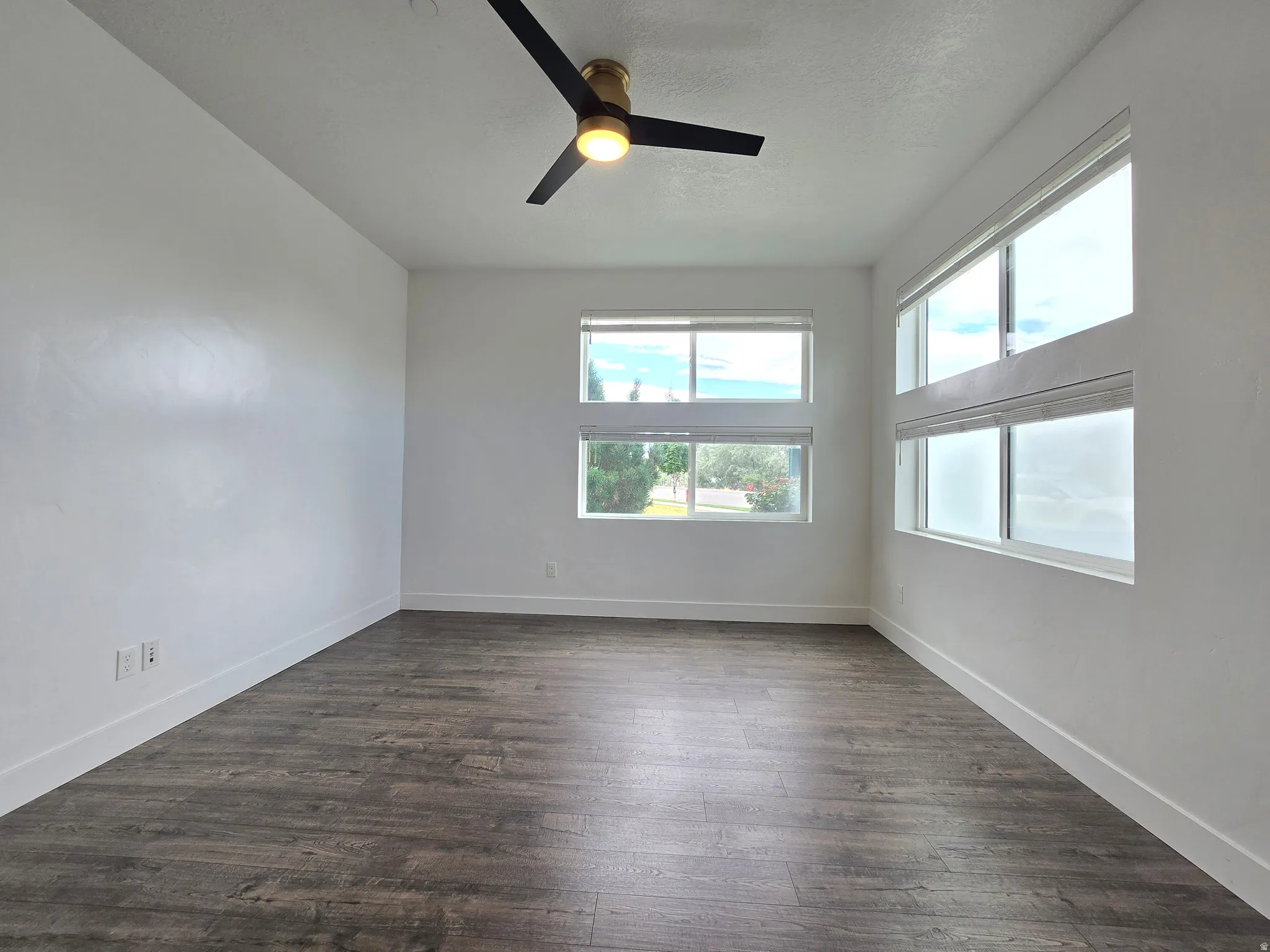 Spacious room featuring extensive wood-finish flooring, multiple windows with white blinds, a contemporary ceiling fan with integrated lighting, and crisp white walls