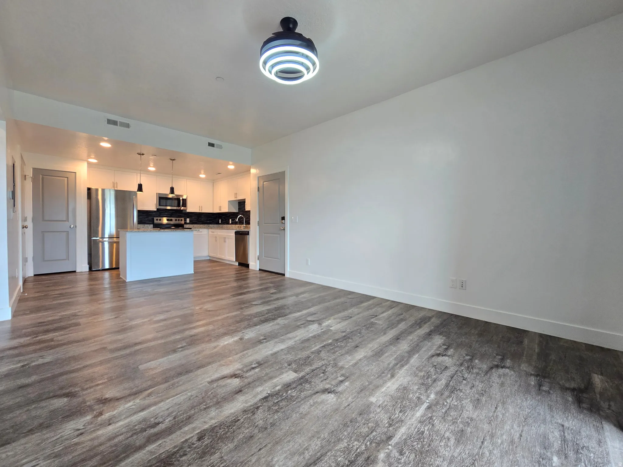 Open-concept living area featuring wood-finish flooring, a contemporary ceiling light fixture, and a kitchen with white cabinetry, stainless steel appliances, and an island