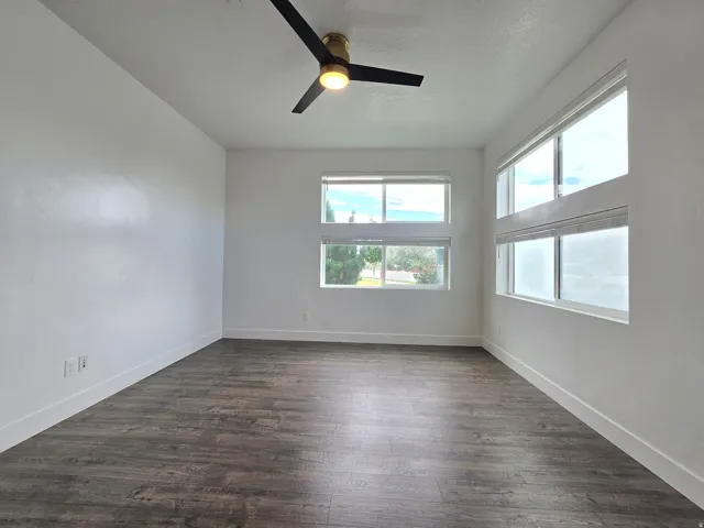 Spacious room featuring extensive wood-finish flooring, multiple windows with white blinds, a contemporary ceiling fan with integrated lighting, and crisp white walls