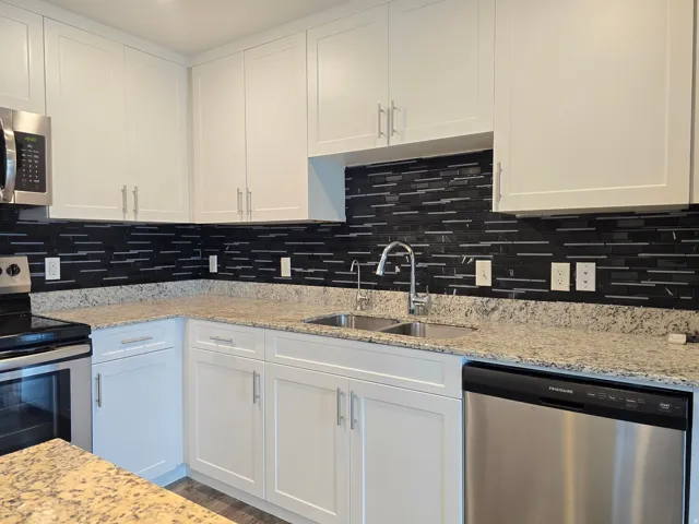 Modern kitchen featuring white shaker cabinetry, stainless steel appliances, speckled stone countertops, a dark mosaic tile backsplash, and a double basin sink with a gooseneck faucet