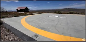 Concrete helipad with yellow marking, featuring a distant cabin with a red roof, set against a backdrop of natural terrain and open sky