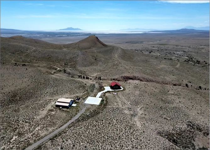 Expansive desert landscape featuring a primary residence with a red roof, an additional outbuilding, a concrete pad, and a gravel driveway, all surrounded by arid terrain and distant mountains under a clear sky