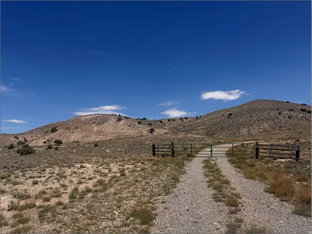 Gravel drive with metal gate opening to expansive land with arid vegetation and distant hills