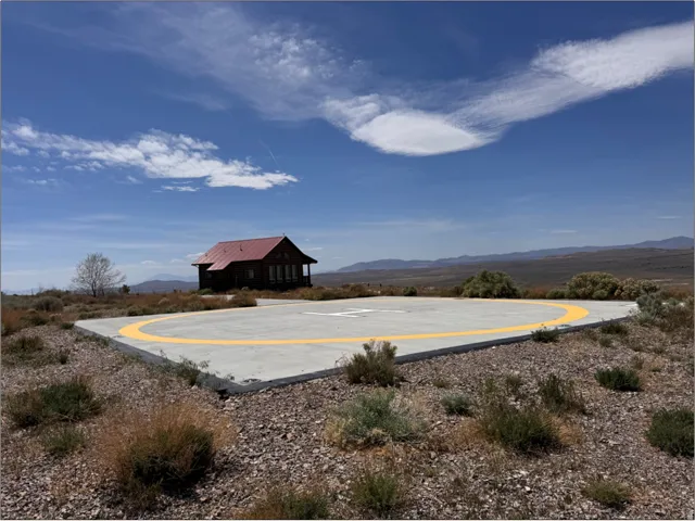 Concrete helipad with yellow perimeter markings positioned on a gravel landscape, featuring a distant cabin with a red roof and covered porch