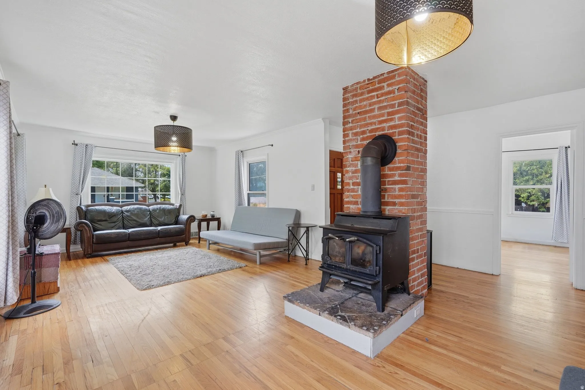 Living area featuring hardwood flooring, a prominent brick column, a black wood-burning stove with a stone hearth, white walls, and a large window