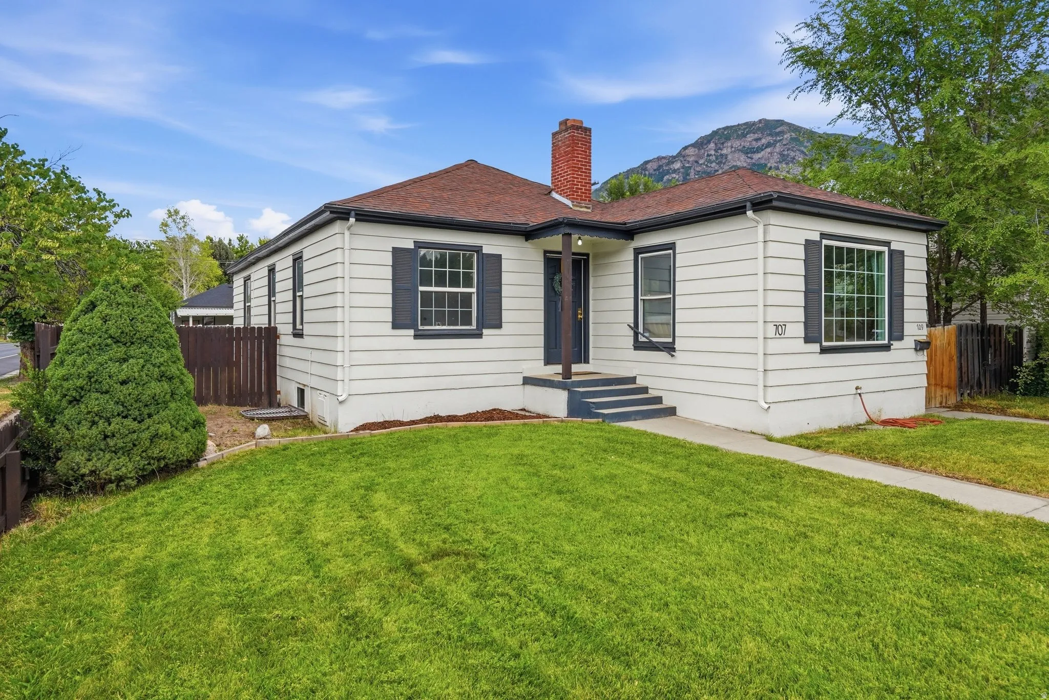 White siding exterior featuring a dark brown shingled roof, dark shutters, red brick chimney, and a landscaped front lawn
