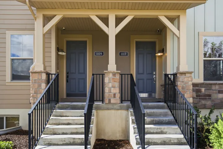 Dual entryways featuring paneled doors, stone-clad pillars, concrete steps with black metal railings, and overhead lighting fixtures