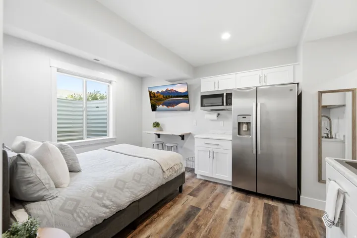 Compact living space featuring wood-finish flooring, a full-size window with a window well, a built-in folding table, white cabinetry with a built-in microwave, and a stainless steel refrigerator
