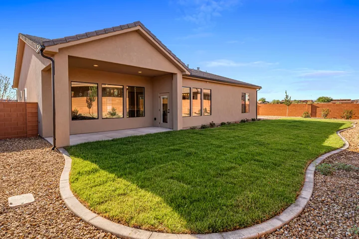 Expansive grassy backyard featuring a covered patio, stucco exterior, multiple large windows, a full-lite exterior door, and gravel landscaping