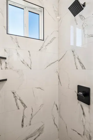 Shower featuring large format white and gray veined wall tiles, a black rectangular rainfall showerhead, and a black square shower control