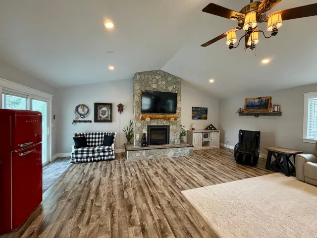 Spacious living area featuring a stone-clad fireplace with a natural wood mantel, wood-finish flooring, vaulted ceilings, recessed lighting, and a ceiling fan with integrated lighting