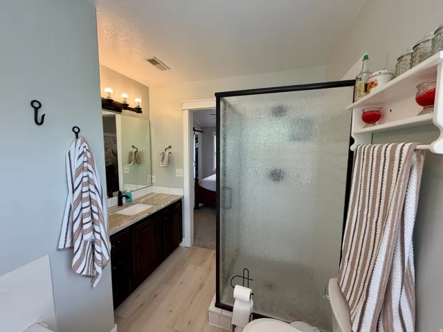Bathroom featuring a shower with a textured glass enclosure, a vanity with a light-toned countertop and dark wood-finish cabinetry, wood-finish flooring, and a wall-mounted light fixture with three bulbs