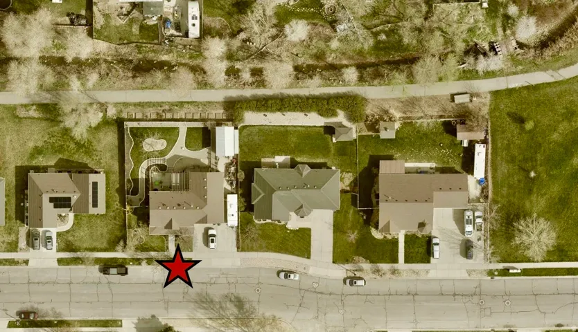 Aerial perspective of a property featuring a brown-roofed residence, a paved driveway, and a backyard with a defined patio area and landscaped elements