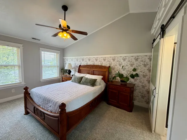 Vaulted ceiling bedroom featuring patterned wallpaper, a ceiling fan with integrated lighting, twin windows with blinds, and a sliding barn door with frosted glass
