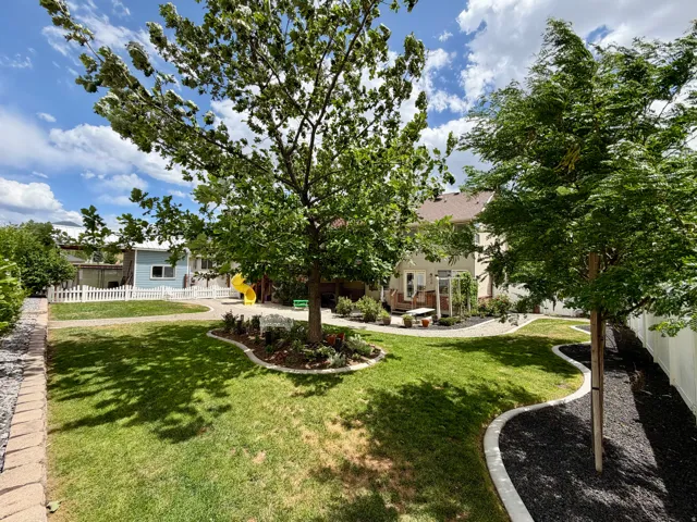 Spacious backyard featuring a manicured lawn, established shade tree, decorative garden beds with concrete curbing, and a white picket fence enclosing a portion of the yard