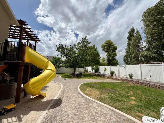 Expansive outdoor area featuring a built-in play structure with a slide, gravel pathways, a manicured lawn, and a white privacy fence